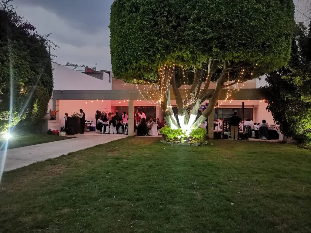 Jardín con pasto y un gran árbol iluminado al anochecer, con el área cubierta y el evento al fondo