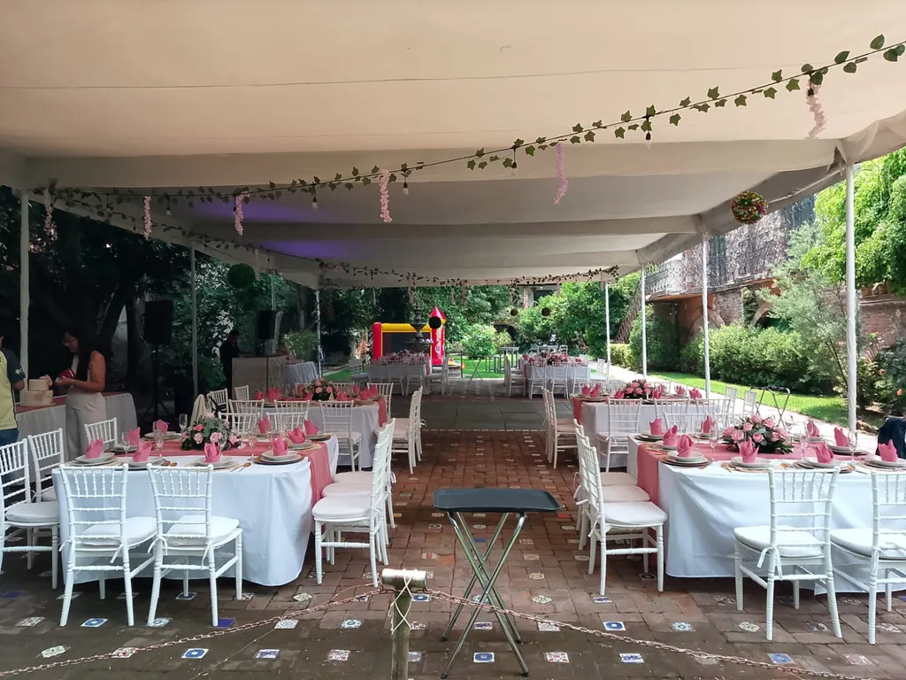 Carpa blanca del Jardín Los Arcos con guías de hojas y focos sobre mesas de mantel blanco con caminos rosas y sillas blancas tipo tiffany, con el jardín al fondo