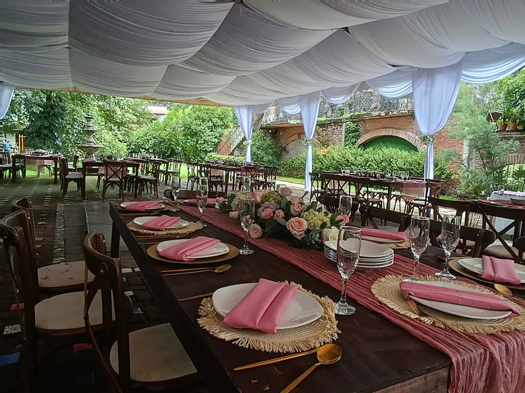 Mesa larga de madera con camino rosa, servilletas rosas y centro de flores bajo un techo de tela blanca drapeada, con muros de ladrillo y arcos al fondo