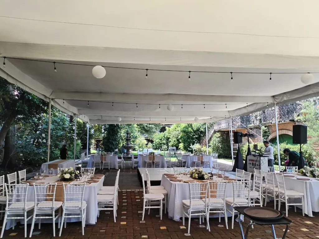 Carpa blanca con series de focos y lámparas de papel sobre mesas de mantel blanco y sillas blancas, con el jardín y la fuente al fondo