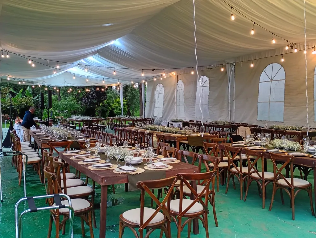 Carpa con techo de tela blanca drapeada, ventanas y series de focos sobre mesas largas de madera con sillas crossback