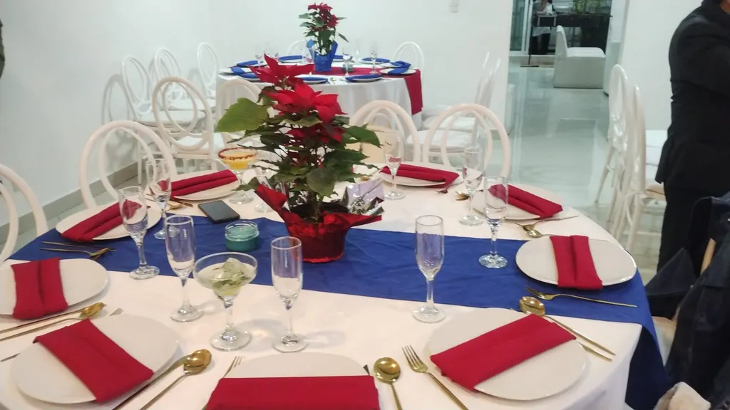 Mesas redondas con mantel blanco y camino azul, servilletas rojas, copas y centros de nochebuena, con sillas blancas de respaldo ovalado