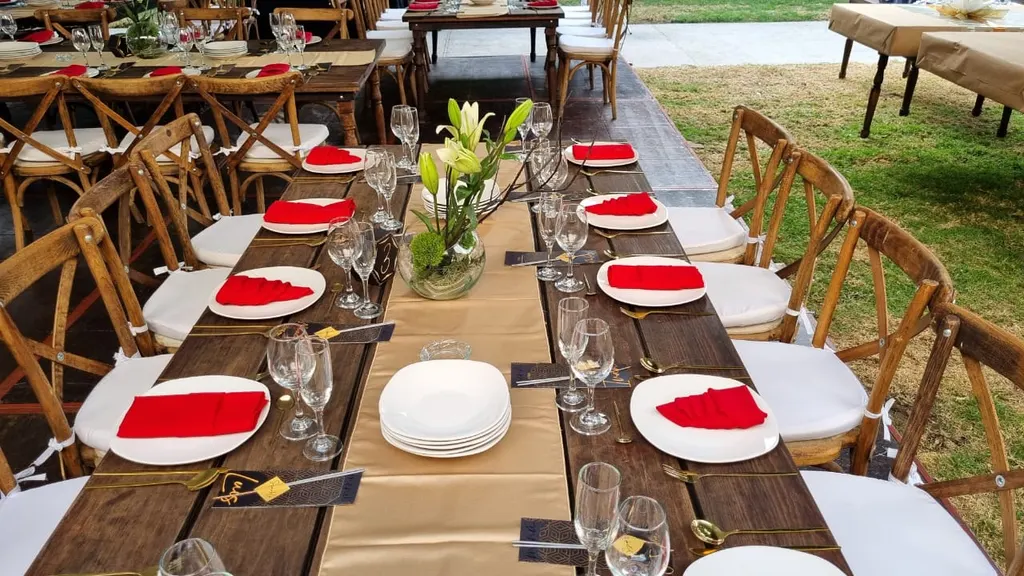 Mesa larga de madera con camino dorado, servilletas rojas, platos blancos, copas y centro de lirios, con sillas crossback de madera sobre el pasto