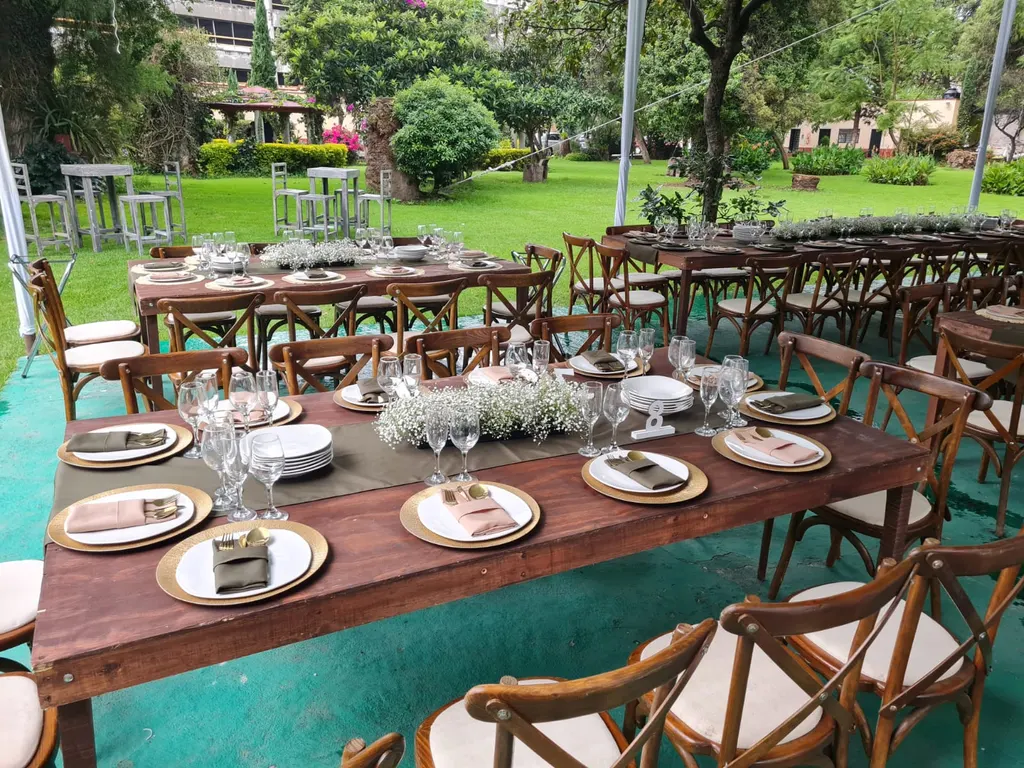 Mesas largas de madera con bajo platos dorados y centros de flores blancas en una carpa con alfombra verde, junto al jardín con pasto y árboles