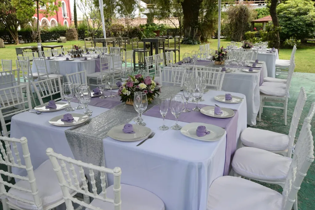 Mesas con mantel blanco, caminos lilas y sillas tiffany sobre el pasto del Jardín Don Bosco, con un edificio de ladrillo rojo al fondo
