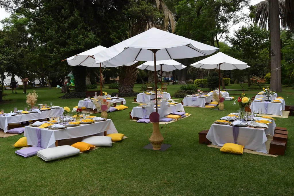 Montaje lounge sobre el pasto con mesas bajas de mantel blanco, cojines amarillos, lilas y blancos y sombrillas blancas