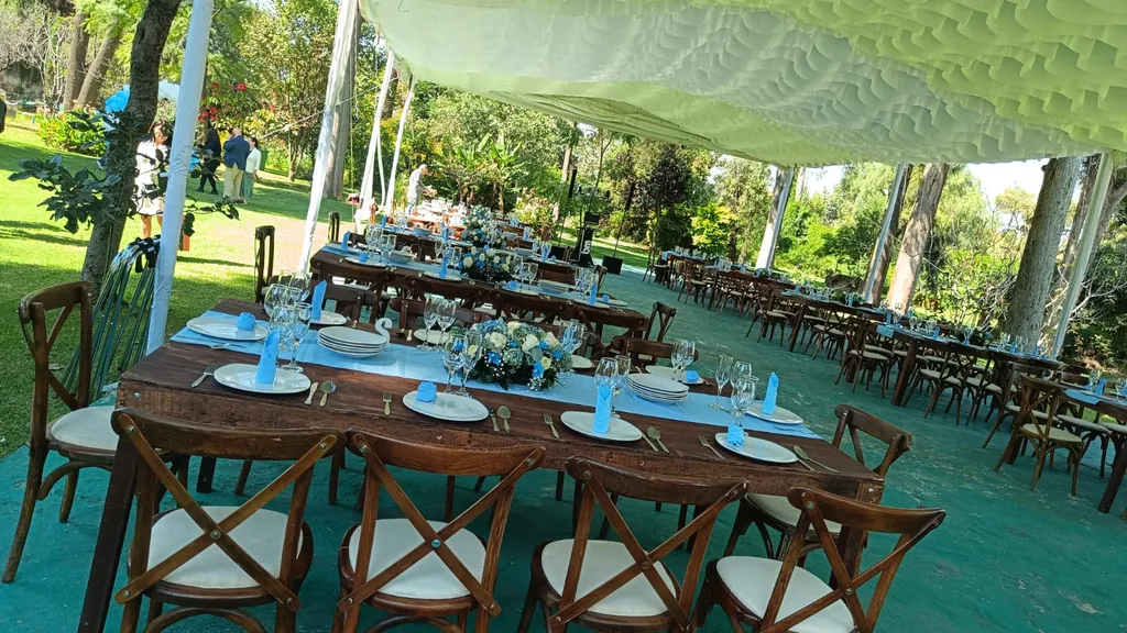 Mesas largas de madera con caminos y servilletas azules y centros de flores blancas y azules bajo una carpa blanca, con sillas crossback y árboles alrededor