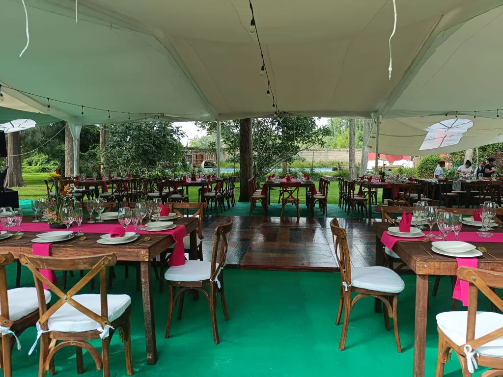 Carpa blanca con series de focos sobre mesas de madera con caminos fucsia, sillas crossback y una pista de madera al centro