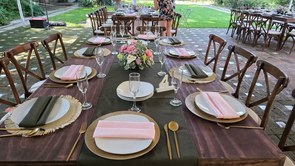 Mesa larga de madera con camino verde olivo, servilletas rosas y verdes, bajo platos de fibra, cubiertos dorados y centro de flores rosas, con sillas crossback