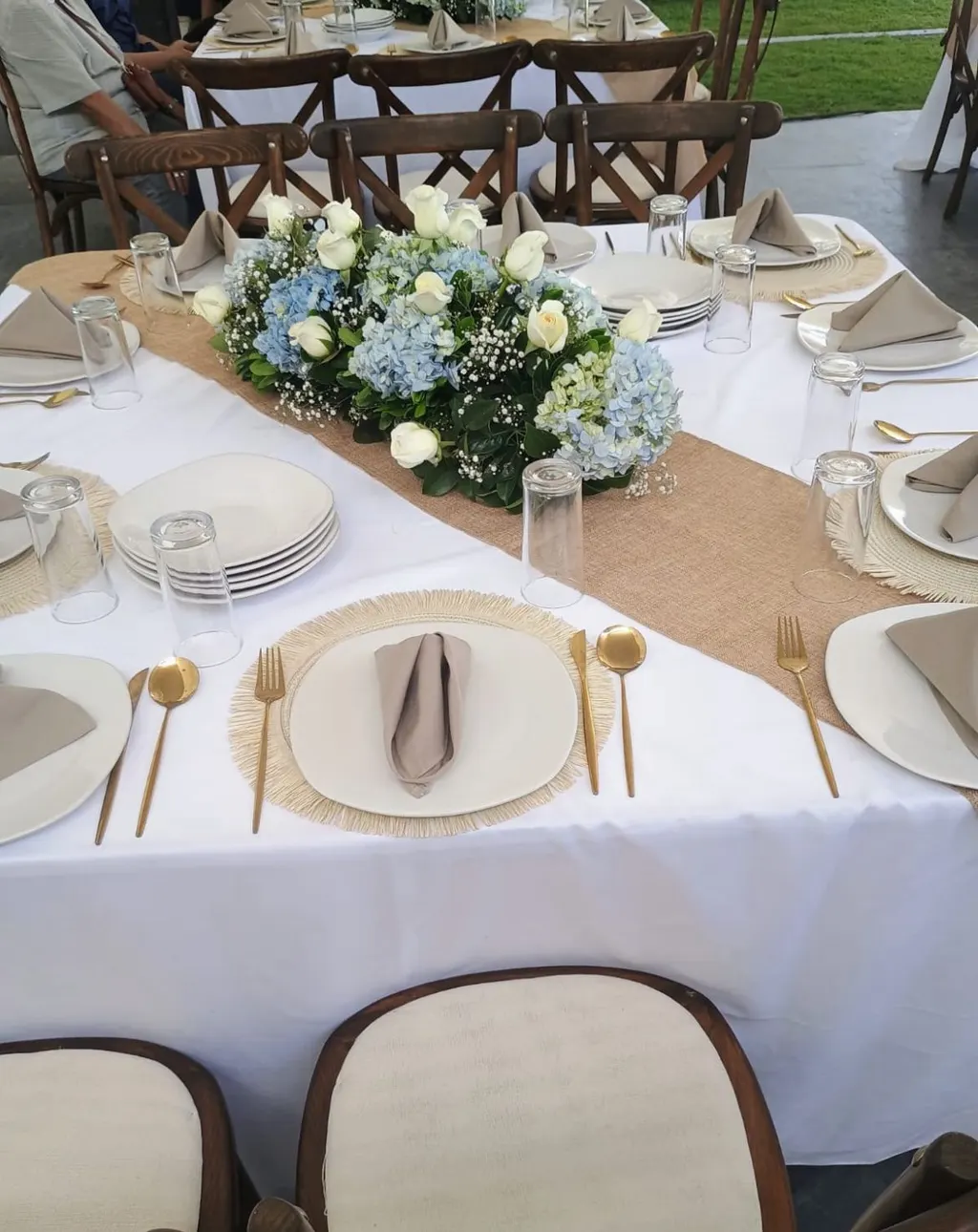 Mesa con mantel blanco, camino de yute, centro de rosas blancas e hortensias azules, platos blancos, cubiertos dorados y sillas de respaldo acolchado